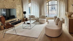 A living room with a large flat-screen TV, a wooden coffee table, a sofa, and two armchairs. There is a window with white curtains and a view of the street outside.