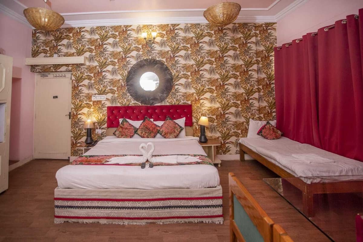 A hotel room with a large bed, a smaller bed, a mirror, a red curtain, and a wooden bench.