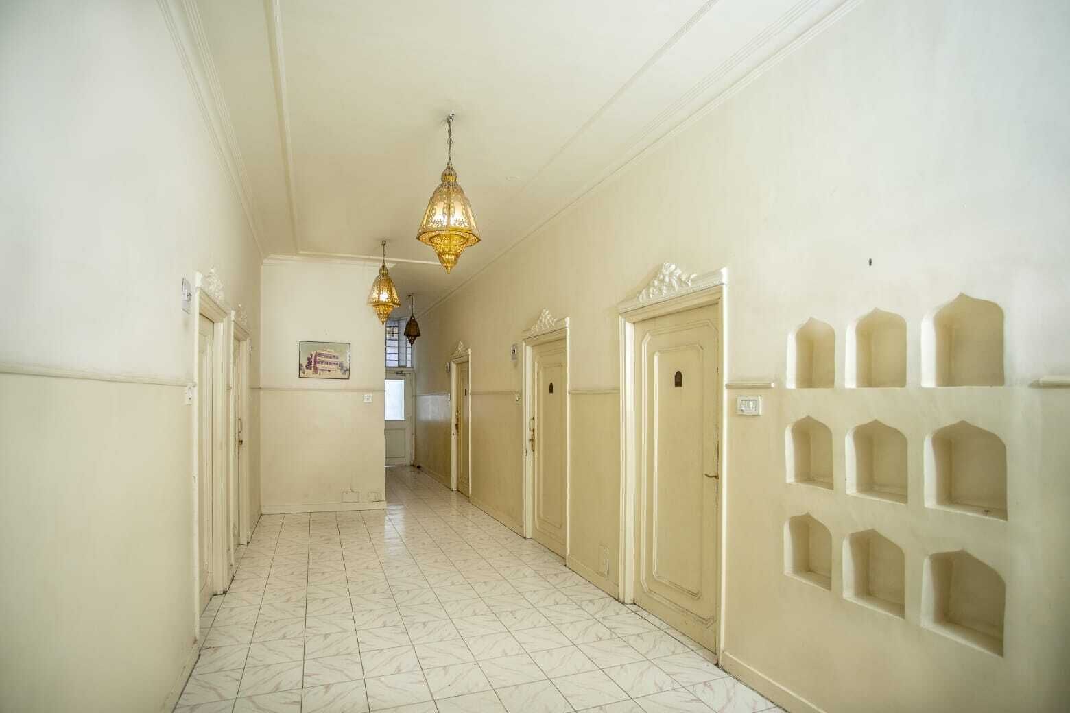 A hallway with white tiled floors and cream-colored walls, featuring doors with decorative moldings and a series of small, recessed windows on the right wall.