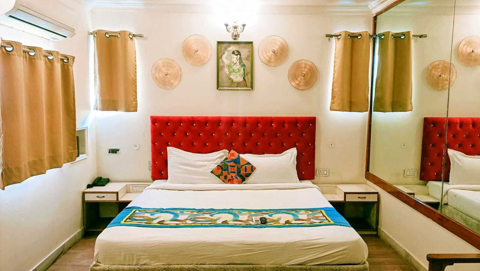 Een hotelkamer met een rode, gestopte hoofdeinde, een bed met witte lakens, een kleurrijk plaid en een grote spiegel aan de muur.