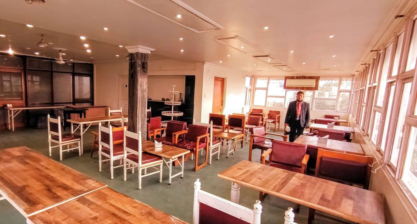A spacious restaurant with wooden tables and red chairs, large windows, and a modern ceiling design.