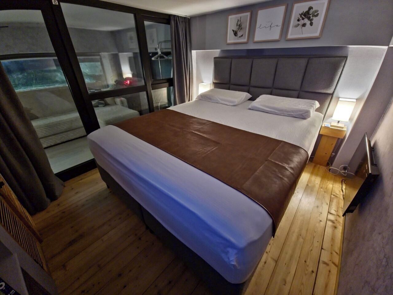 A bedroom with a bed, a window, a bedside table, and a wall with framed pictures.