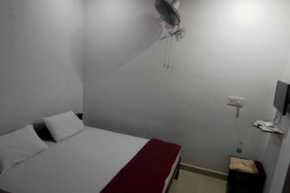 A single bed with a white and red bedspread, a wall-mounted fan, a television, and a small trash bin.
