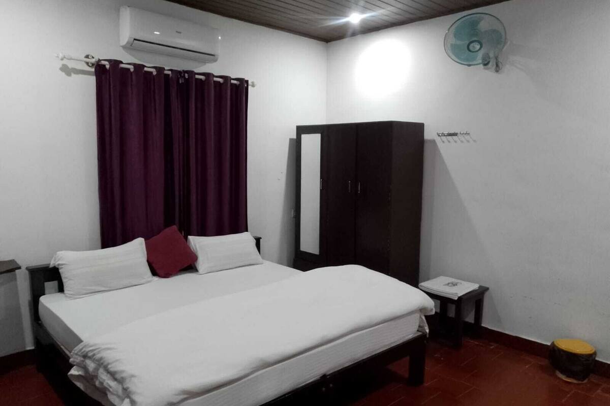 A bedroom with a bed, a wardrobe, an air conditioner, a fan, and a small table with a book.
