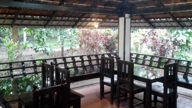 An outdoor covered seating area with wooden tables and chairs, surrounded by a metal railing, and a view of greenery outside. - Humble Nest Coorg (Somvarpet)
