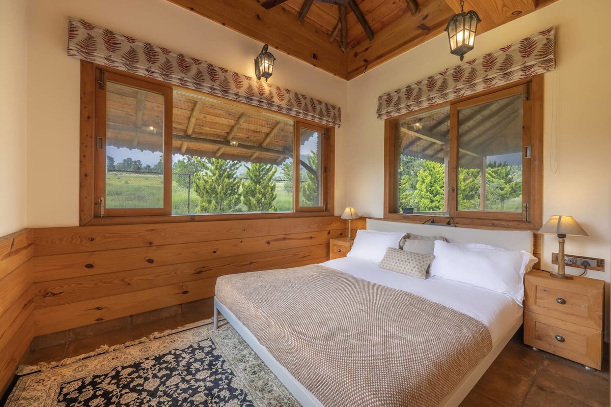 Un dormitorio con cabecera de madera, una cama grande, dos lámparas de noche, una mesita de noche y una ventana con vistas a la vegetación.