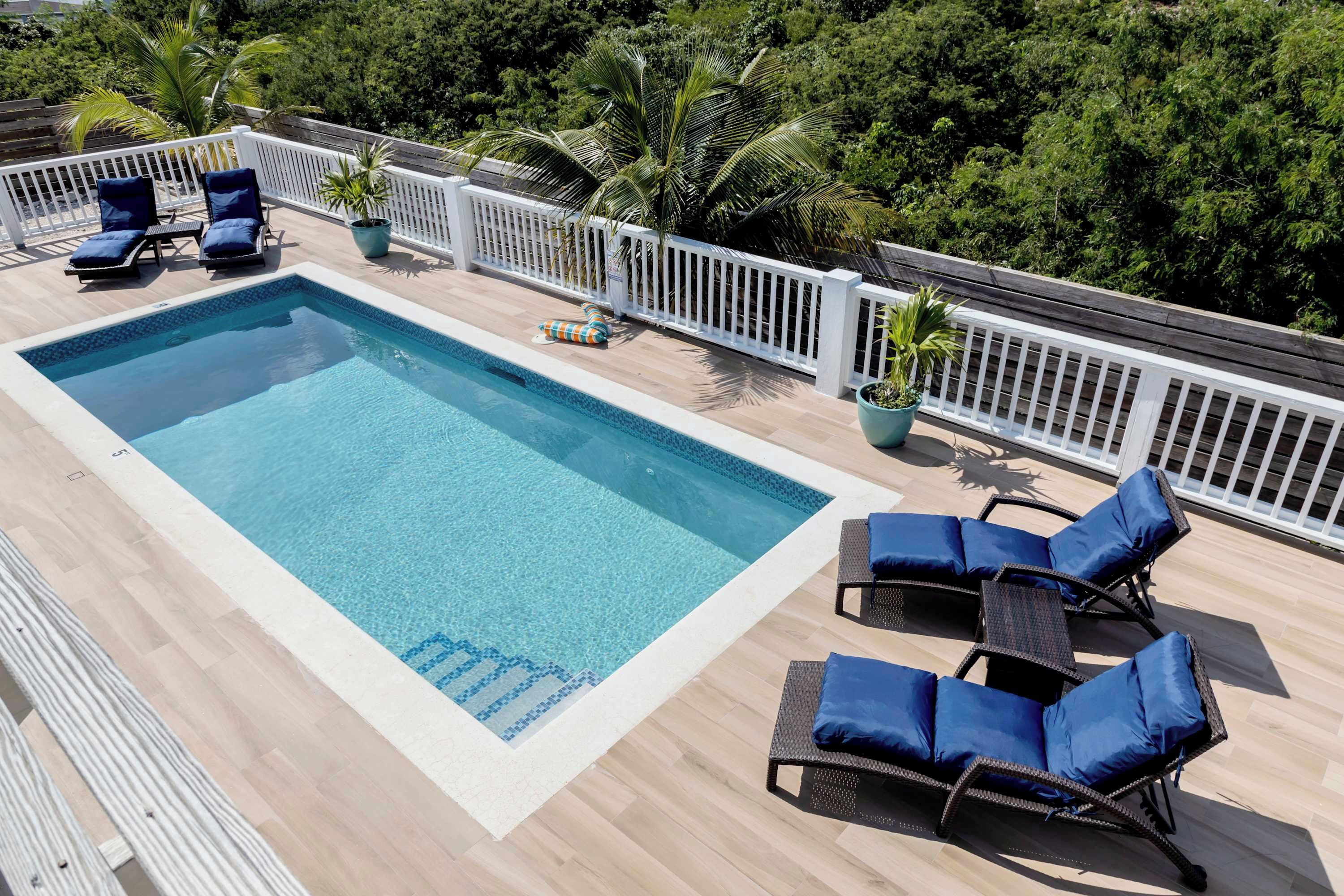 Una piscina rectangular con agua azul, rodeada por un muelle de madera. Hay sillas de descanso con cojines azules y una cerca blanca.