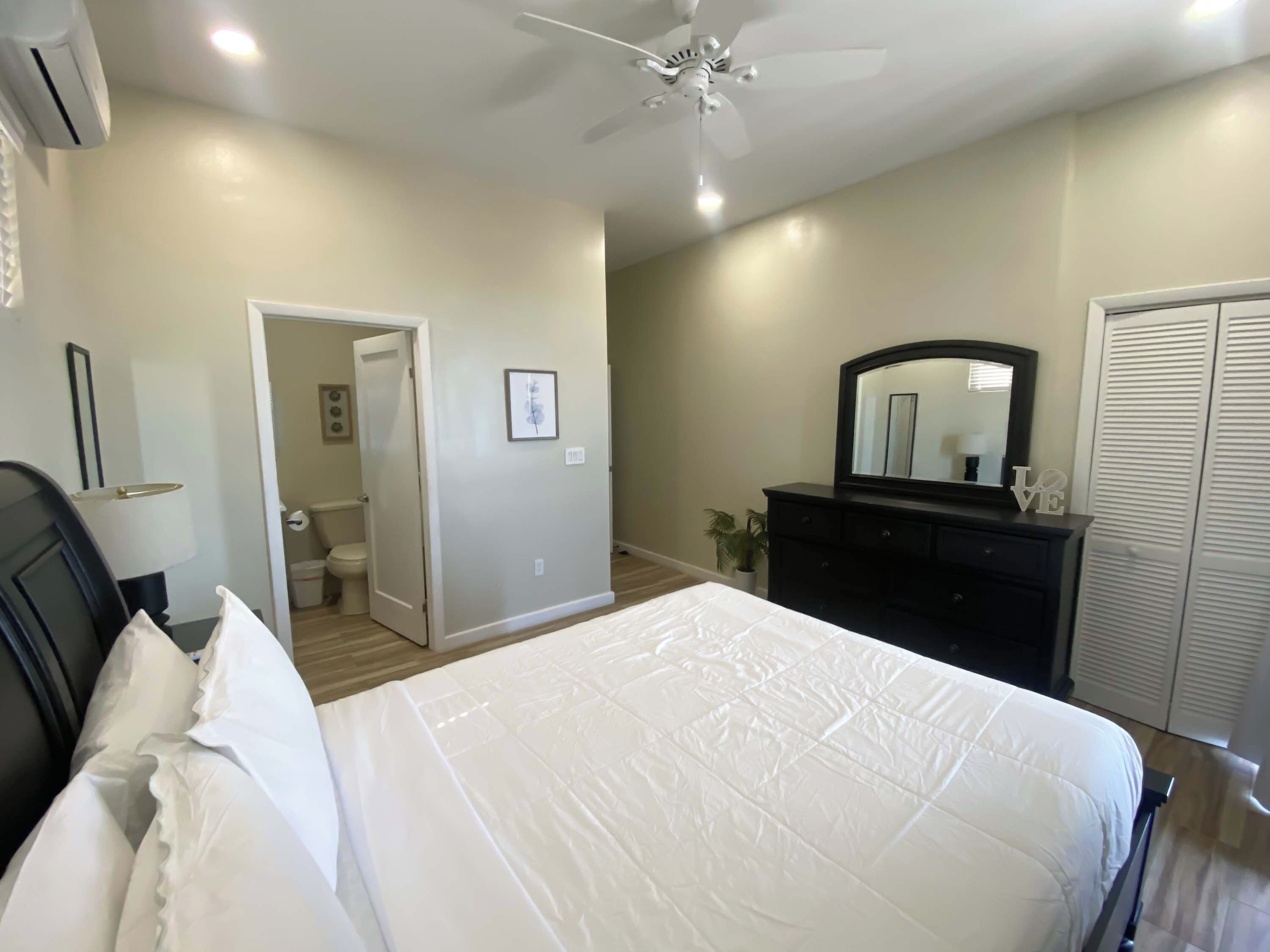 A bedroom with a bed, a dresser, a ceiling fan, and a door leading to a bathroom.