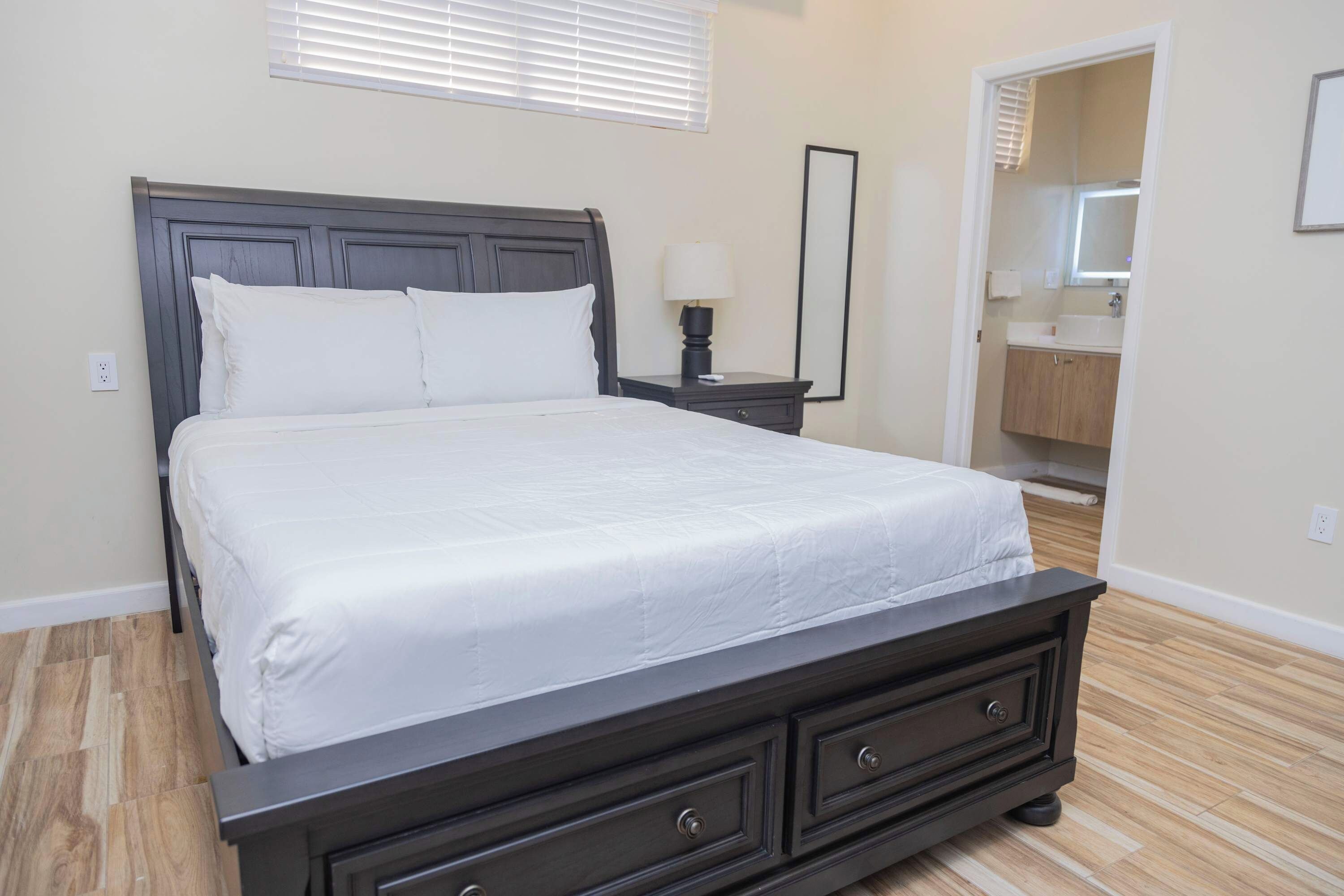 A bedroom with a large bed, a nightstand with a lamp, a dresser, a mirror, and a bathroom visible through an open door.