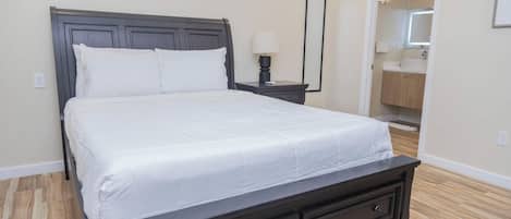 Un dormitorio con una cama grande, una mesita de noche con lámpara, un aparador, un espejo y un baño visible a través de una puerta abierta.