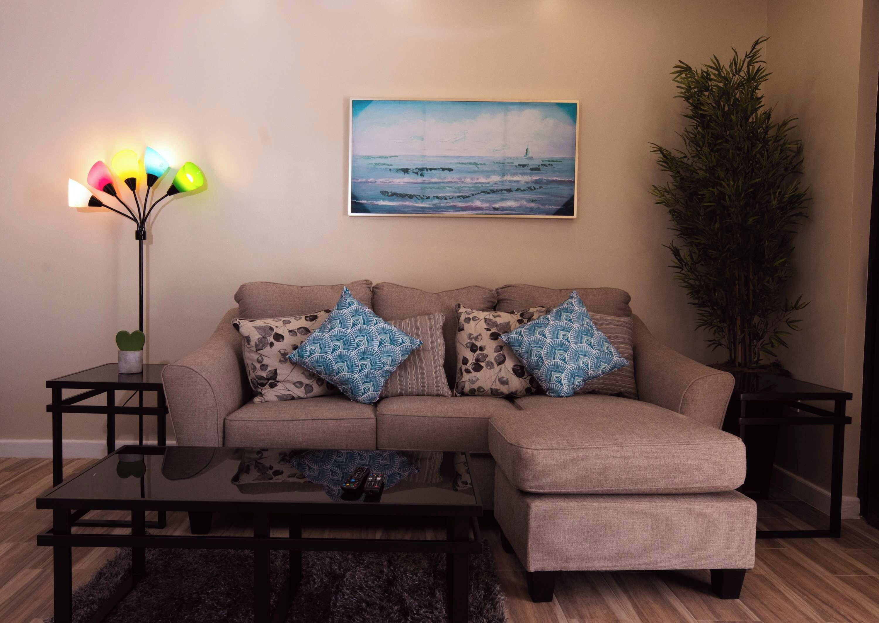Un salon avec un canapé d’angle beige, deux coussins décoratifs à motifs, une table basse en verre, un lampadaire et un tableau représentant un paysage marin.