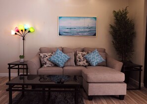 A living room with a beige sectional sofa, two patterned throw pillows, a glass coffee table, a floor lamp, and a painting of a seascape.