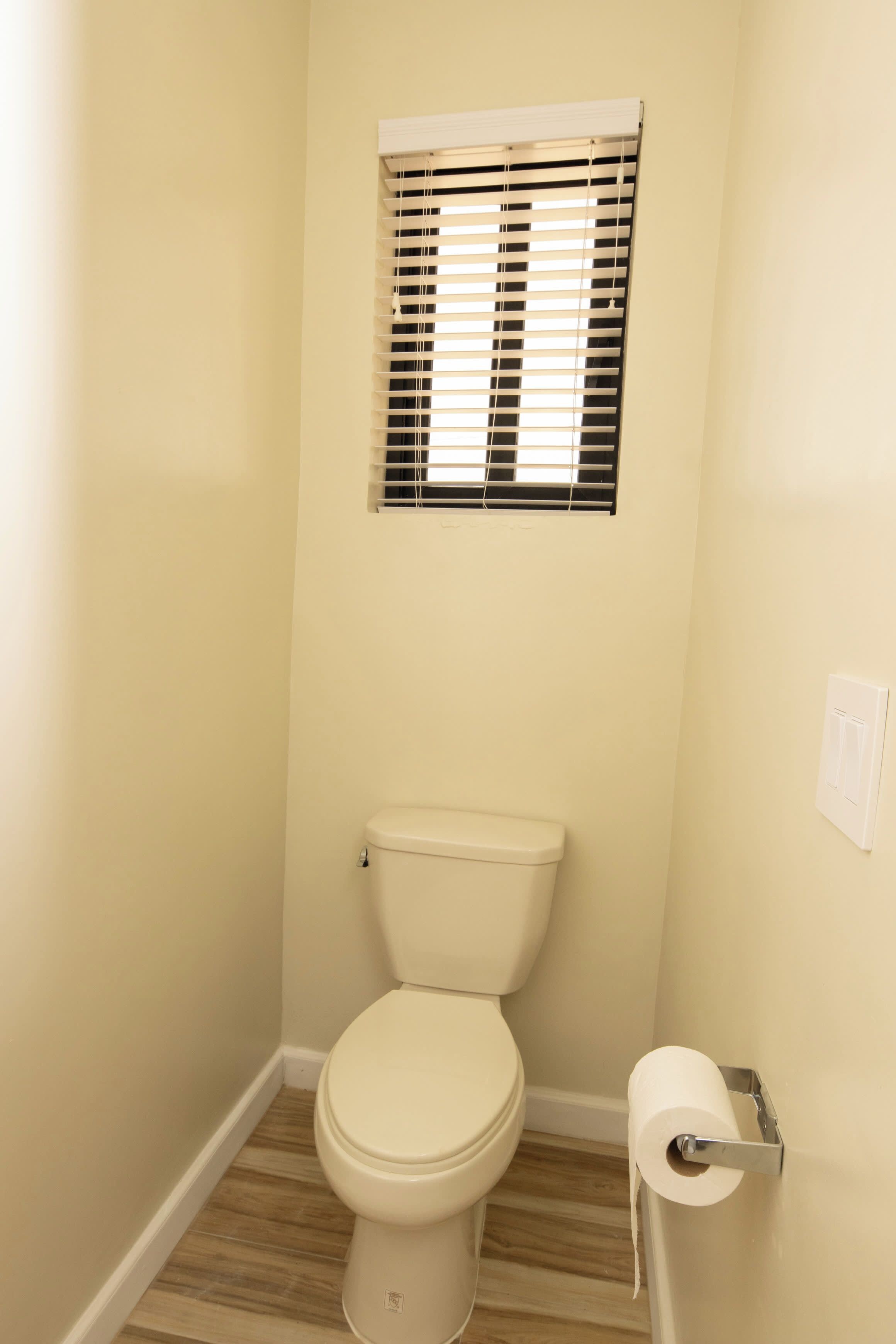 A small, clean bathroom with a toilet, a roll of toilet paper, and a window with blinds.