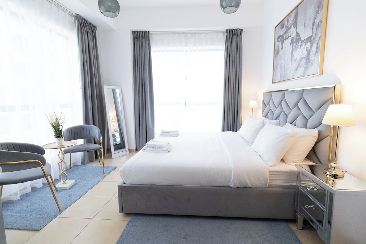 A modern hotel room with a large bed, a desk, a chair, and a large window with curtains.