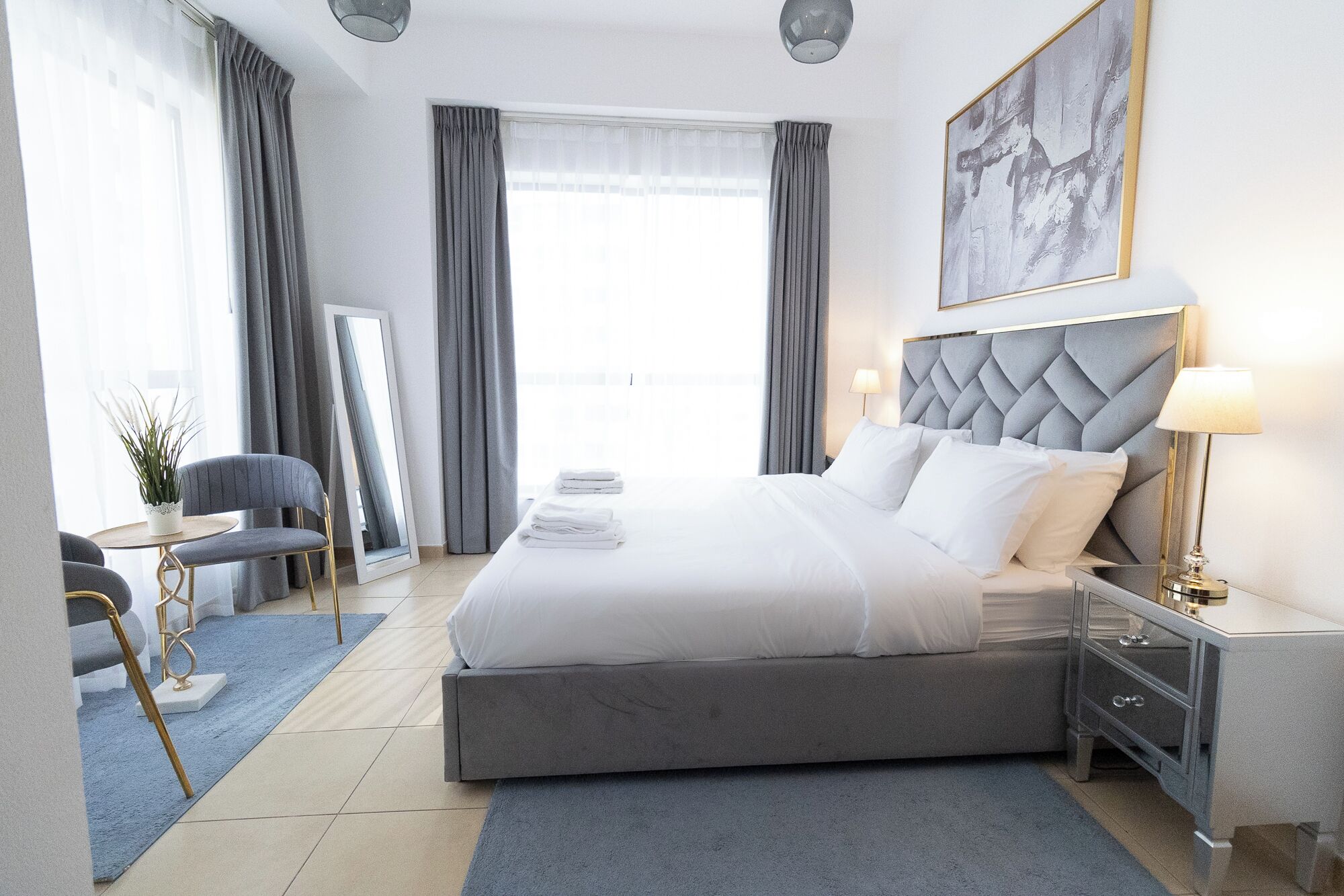 Habitació d'hotel moderna amb un llit gran, una taula de nit amb una làmpada, una cadira, una taula petita amb una planta i un finestral gran amb cortines.