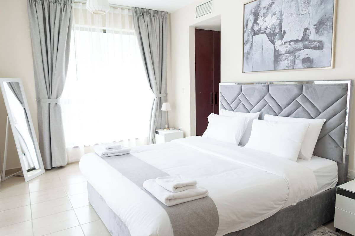 A modern hotel room with a large bed, grey curtains, a painting on the wall, and a white bedside table.