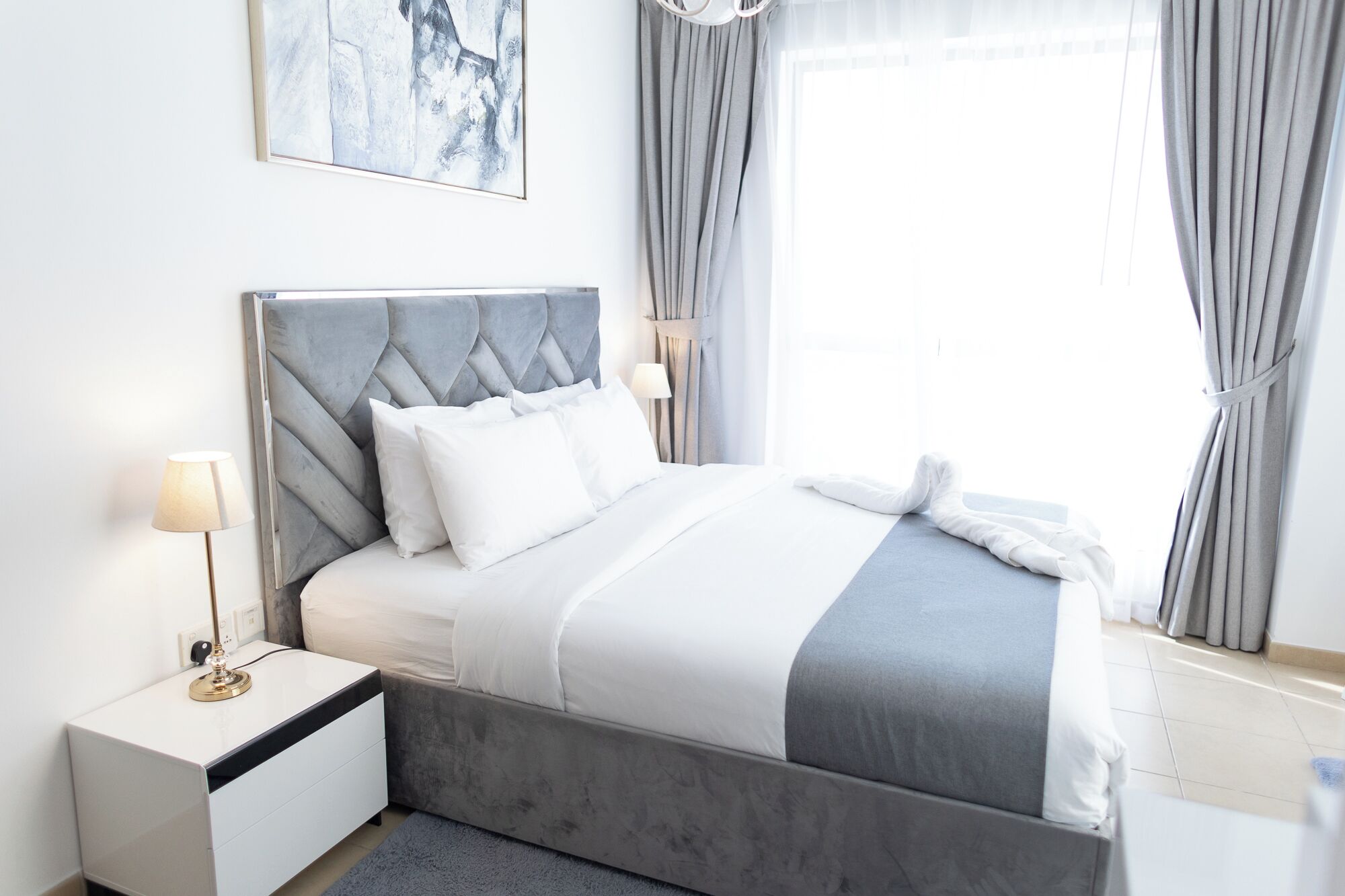 Une chambre à coucher moderne avec une tête de lit rembourrée de couleur grise, du linge de lit blanc et une table de chevet avec une lampe.