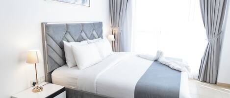 Une chambre à coucher moderne avec une tête de lit rembourrée de couleur grise, du linge de lit blanc et une table de chevet avec une lampe.