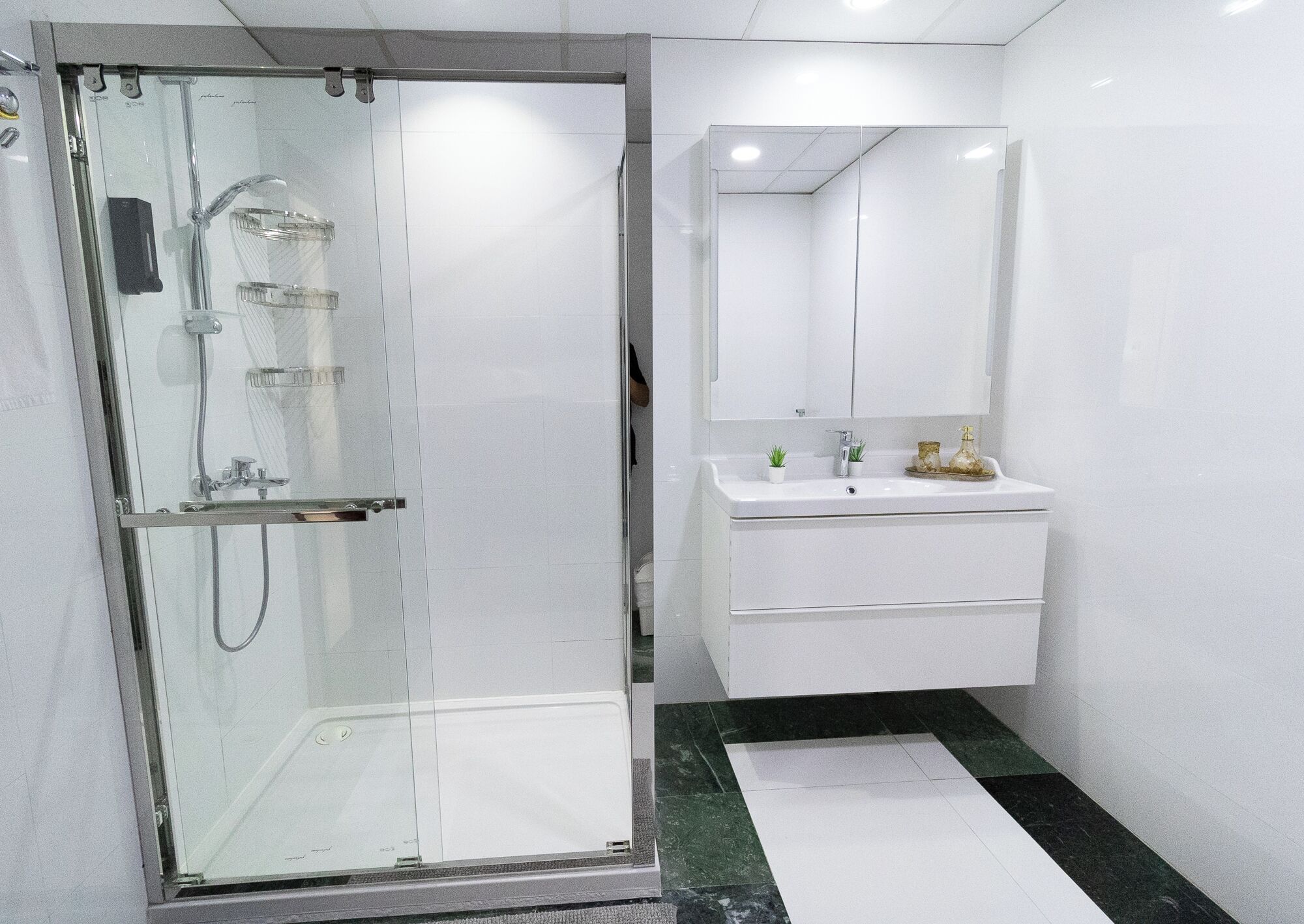 Un baño moderno con ducha envuelta en vidrio, tocador blanco con espejo y piso de baldosas verdes y blancas.