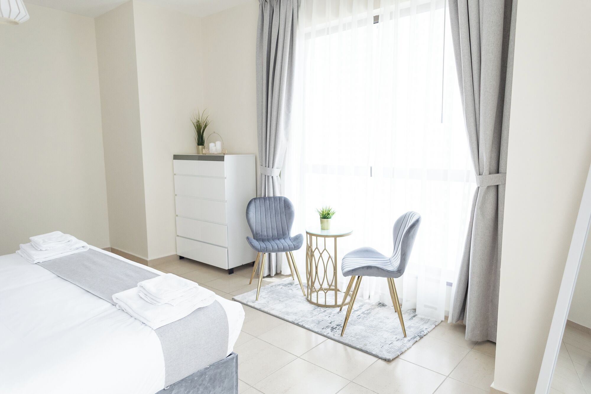 A hotel room with a bed, two chairs, a small table, a dresser, and a window with curtains.