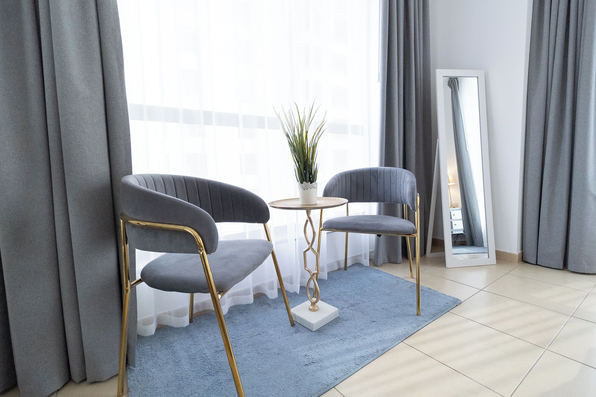 A modern living room with two grey upholstered chairs, a small round table, a potted plant, and a large mirror.