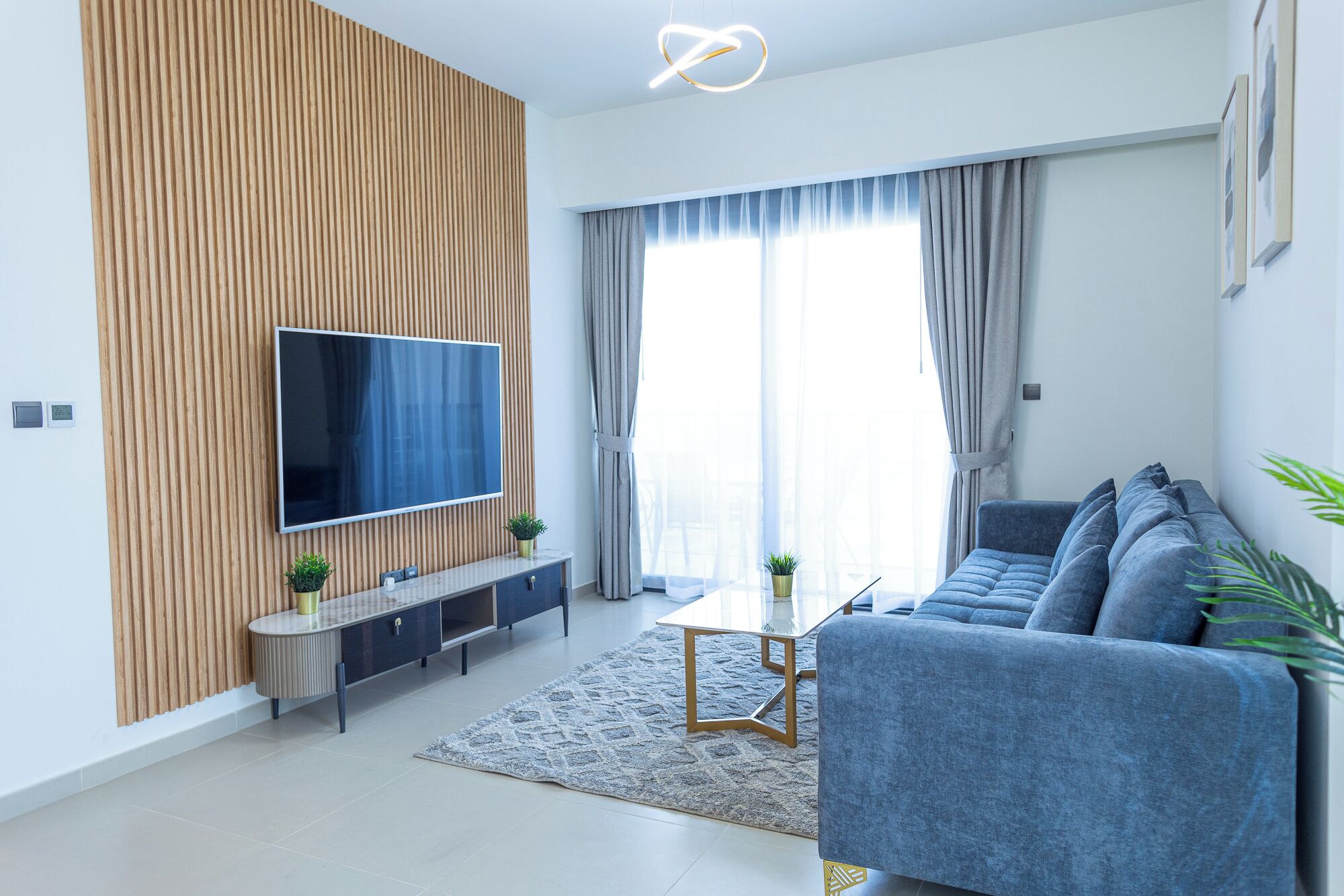 Ein modernes Wohnzimmer mit einem Flachbildfernseher an einer Wand aus Holzpaneelen, einem grauen Sofa, einem Couchtisch und einem kleinen Beistelltisch mit einer Pflanze.