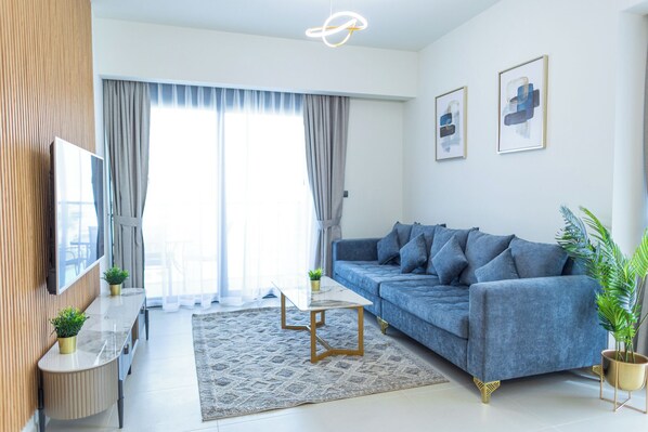 A modern living room with a blue sofa, a glass coffee table, a TV, and a large window with curtains.