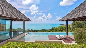 A modern infinity pool with a clear view of the ocean, surrounded by lush greenery and two wooden-roofed structures.