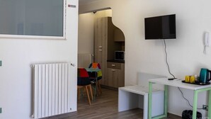 Een compacte kamer met een witte radiator, een aan de muur bevestigde televisie, een klein tafeltje met een waterkoker en een kleurrijke stoel.