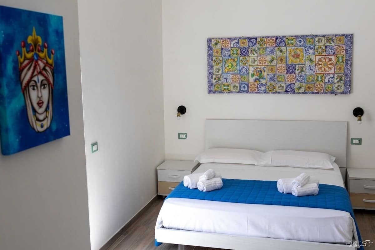 A hotel room with a bed, two bedside tables, a blue bedspread, white pillows, and a colorful tile artwork above the bed.