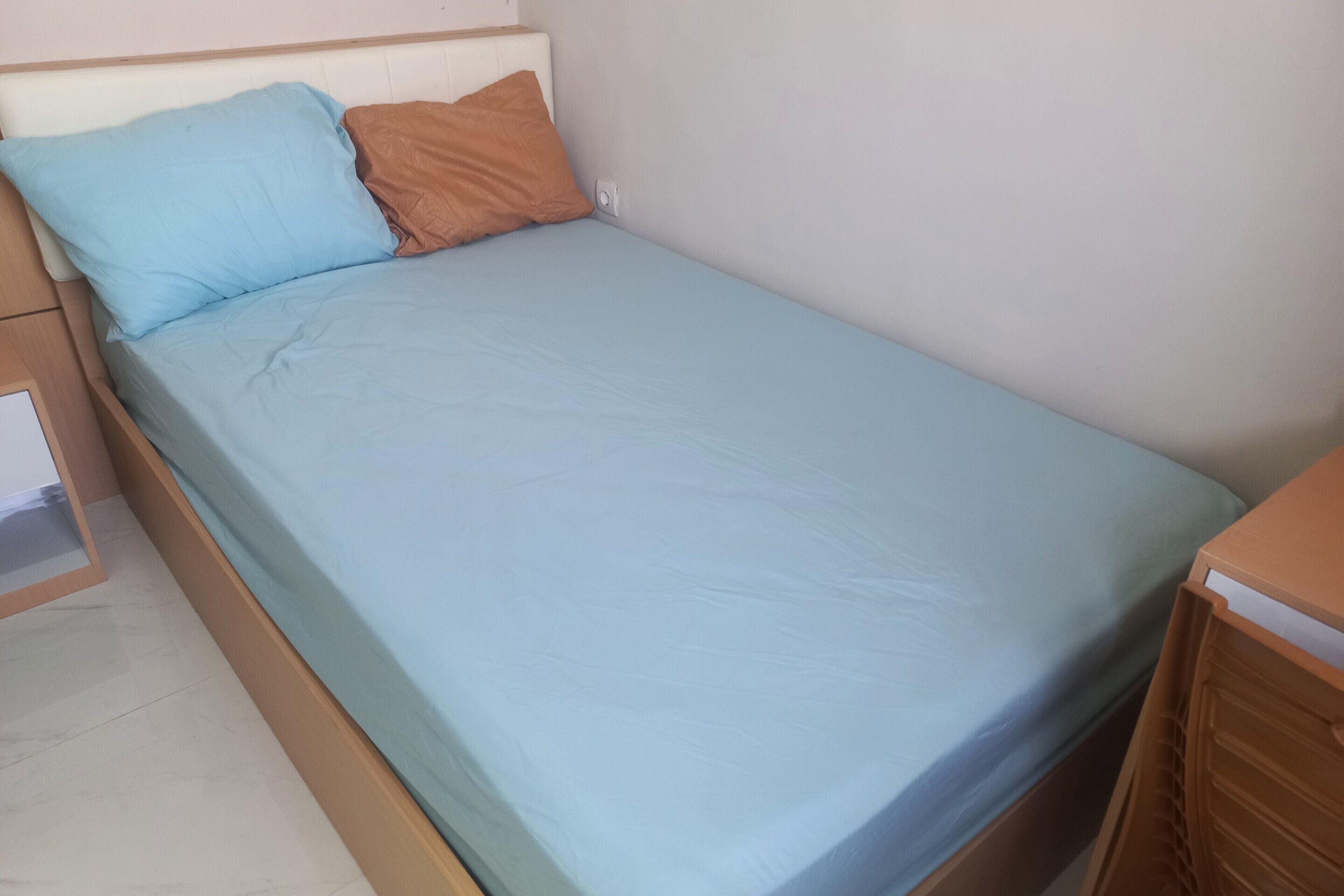 A single bed with a light blue mattress and two pillows, one blue and one brown, in a room with a plain white wall and a wooden nightstand to the left.