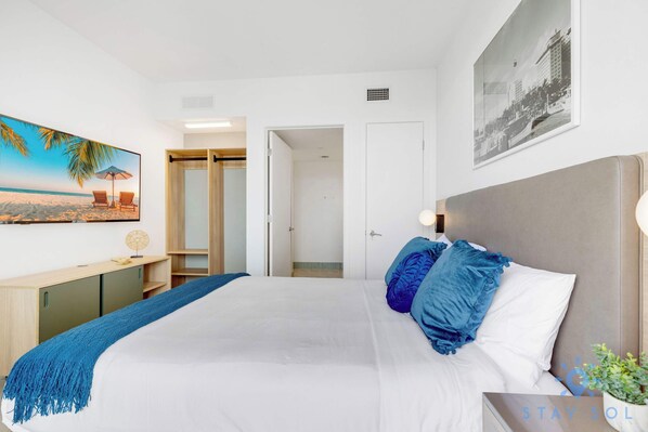 Une chambre à coucher avec un grand lit, une table de chevet, une commode, une lampe fixée au mur, un tableau encadré et une porte donnant sur une autre pièce.