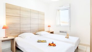A hotel room with two beds, a wooden headboard, bedside lamps, a window with a view, and a toy car on the bed. - Cap Med by Interhome (Le Grau du Roi)