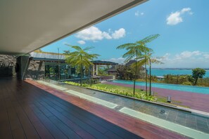 A modern outdoor area with a wooden deck, a glass enclosure, and a view of a pool and ocean. - Beyond The Sea (Jimbaran)