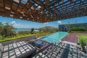 A modern outdoor pool area with a wooden pergola overhead, a seating area with cushions, and a view of the sea. - Beyond The Sea (Jimbaran)