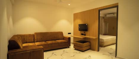 Una sala de estar moderna con un sofá marrón, un televisor de pantalla plana montado en un panel de madera, y una cama en la habitación contigua.