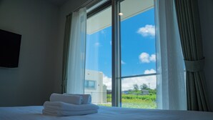 A hotel room with a bed, a window with curtains, and a view of greenery outside. - Porte Bonheur (Miyakojima)