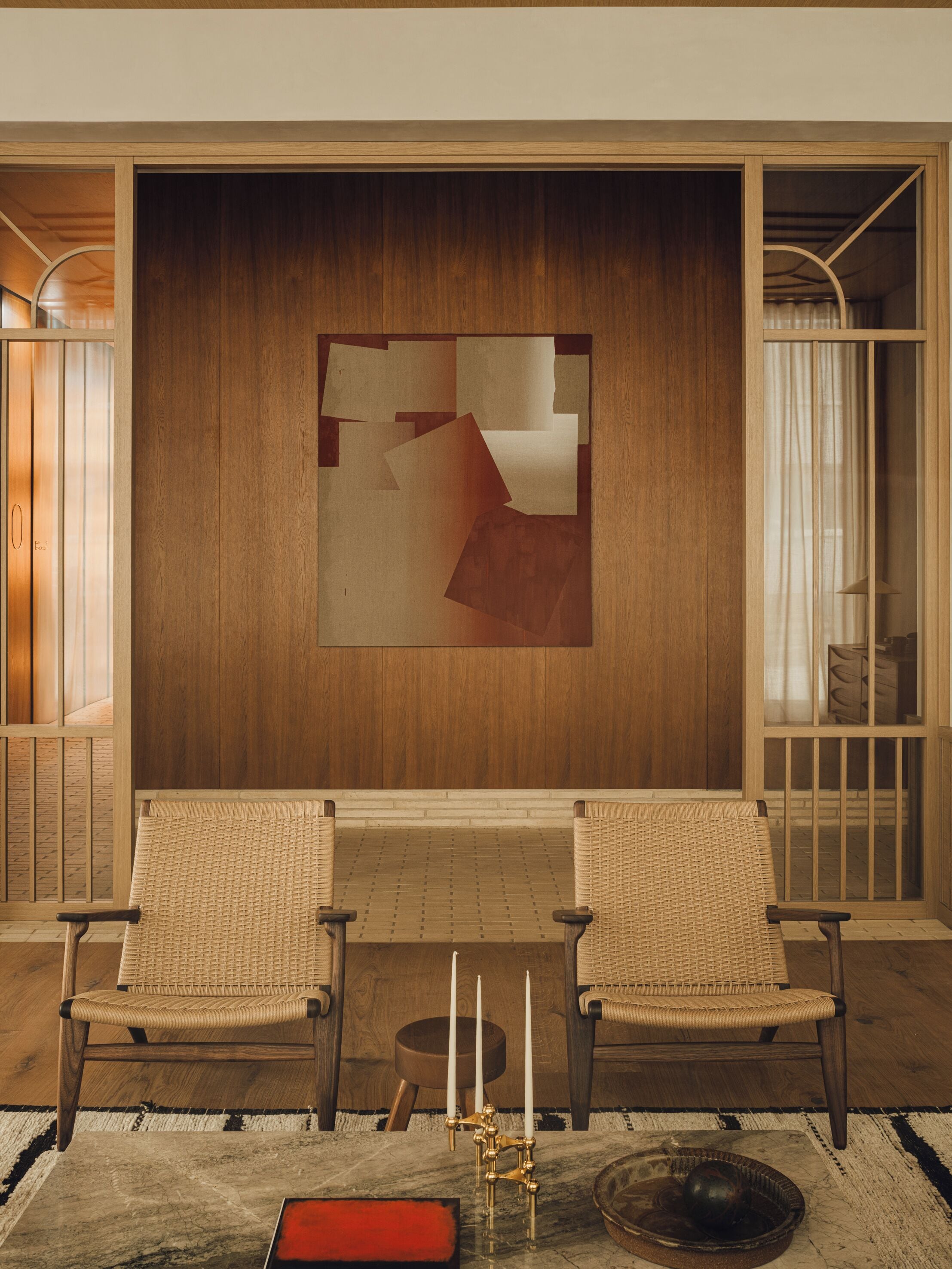 A room with two wicker chairs, a wooden wall with a geometric artwork, a small table with candles, and a window with curtains.