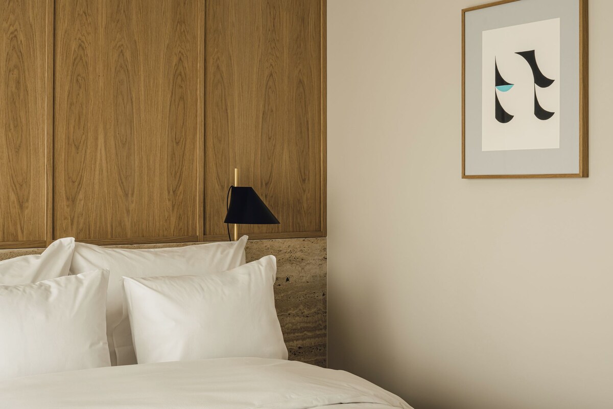 A bedroom with a wooden headboard, a bed with white linens, a black floor lamp, and a framed abstract artwork on the wall.