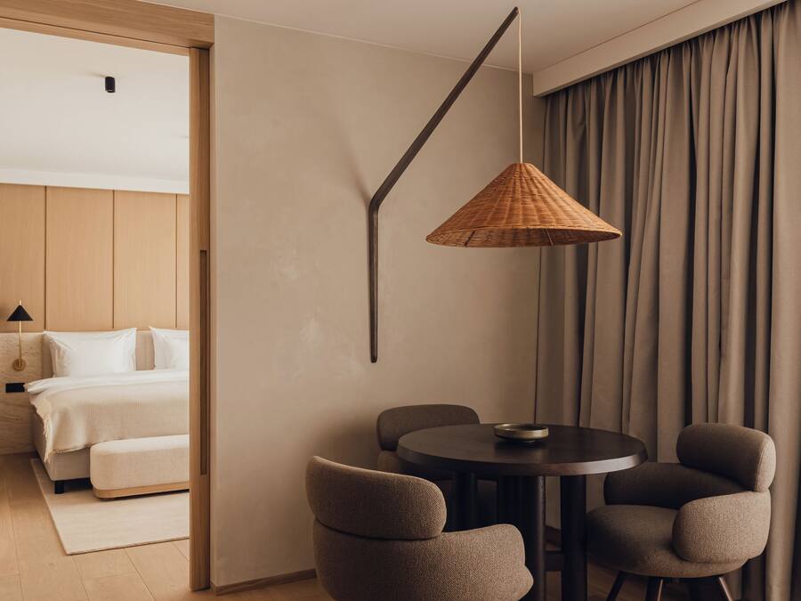 A modern hotel room with a wooden floor, a round dining table with four chairs, a hanging woven light fixture, and a view of a bedroom with a bed and curtains.