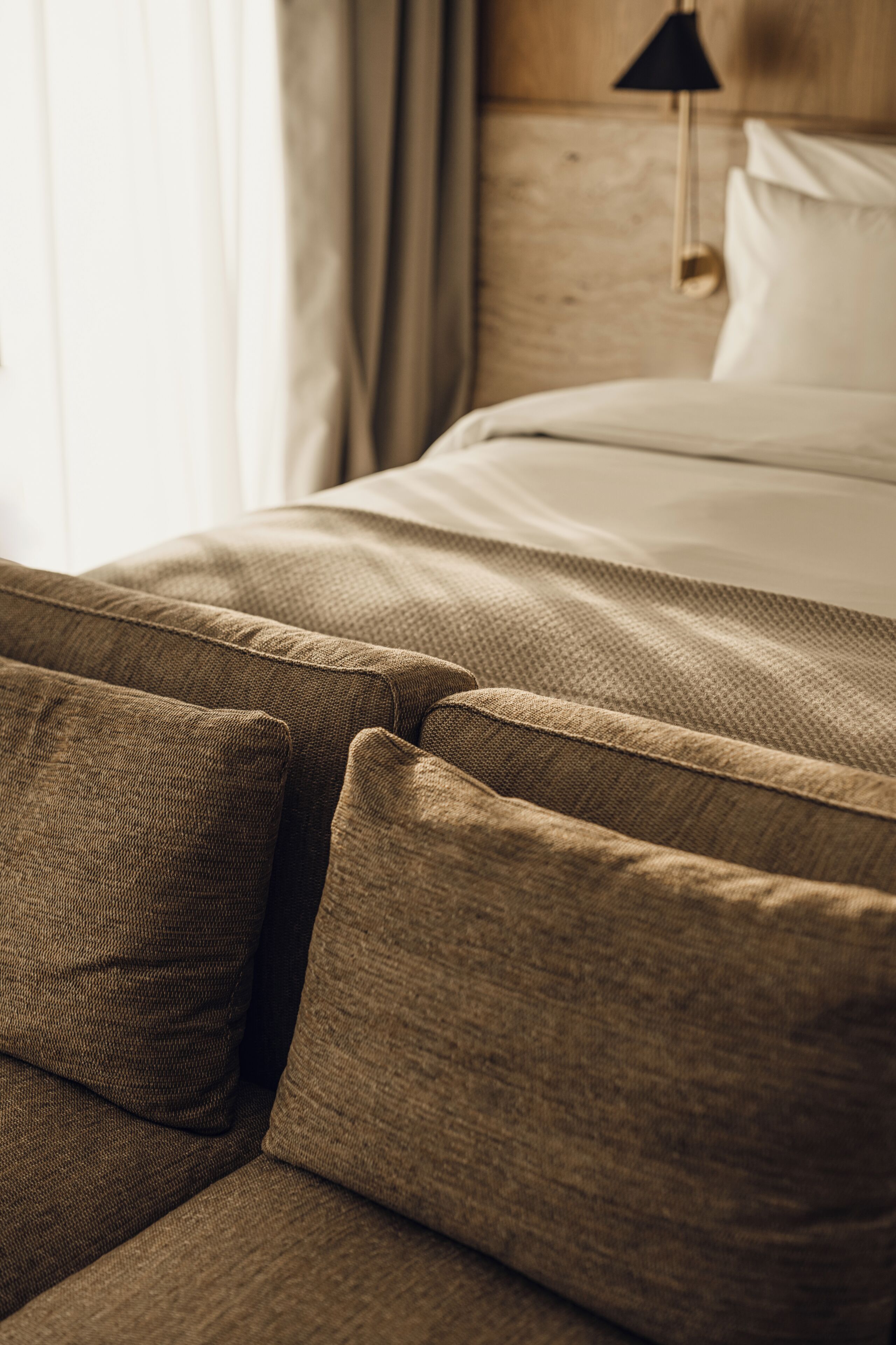 A close-up of a textured brown sofa with a cushion, part of a hotel room with a wooden headboard and a hanging lamp.