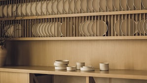 Une étagère en bois avec des assiettes et des bols, une chaise et une table sur laquelle se trouvent un vase et un saladier.