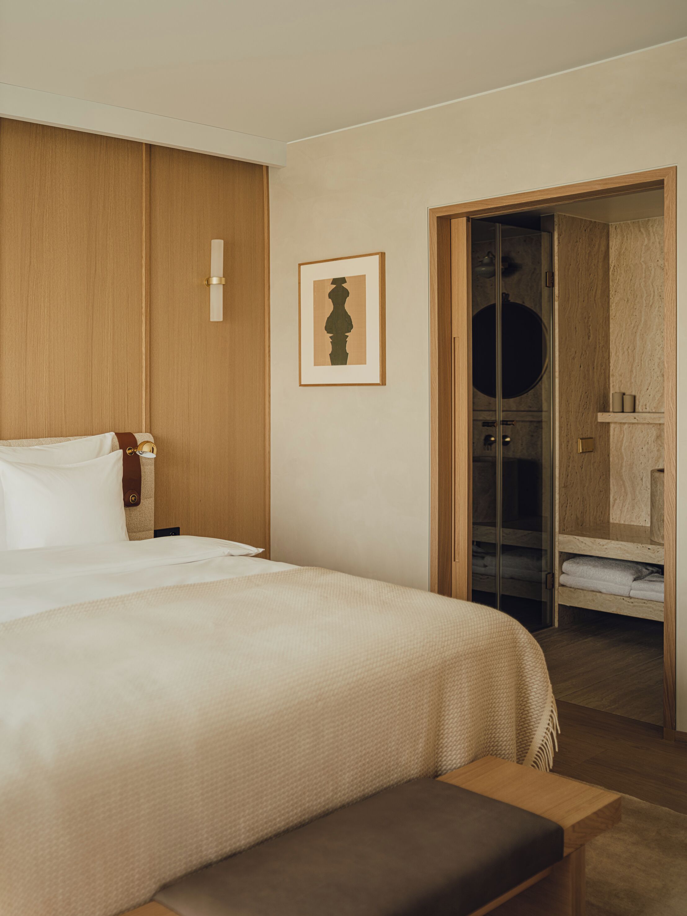 A hotel room with a large bed, a wooden wardrobe, a framed artwork on the wall, and a bathroom visible through an open door.