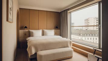 Habitación de hotel moderna con una cama grande, un banco, vista de edificios a través de un ventanal y un cabecero de madera.