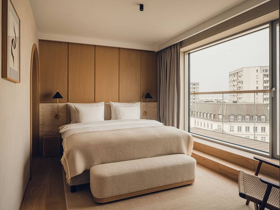 Habitación de hotel moderna con una cama grande, un banco, vista de edificios a través de un ventanal y un cabecero de madera.