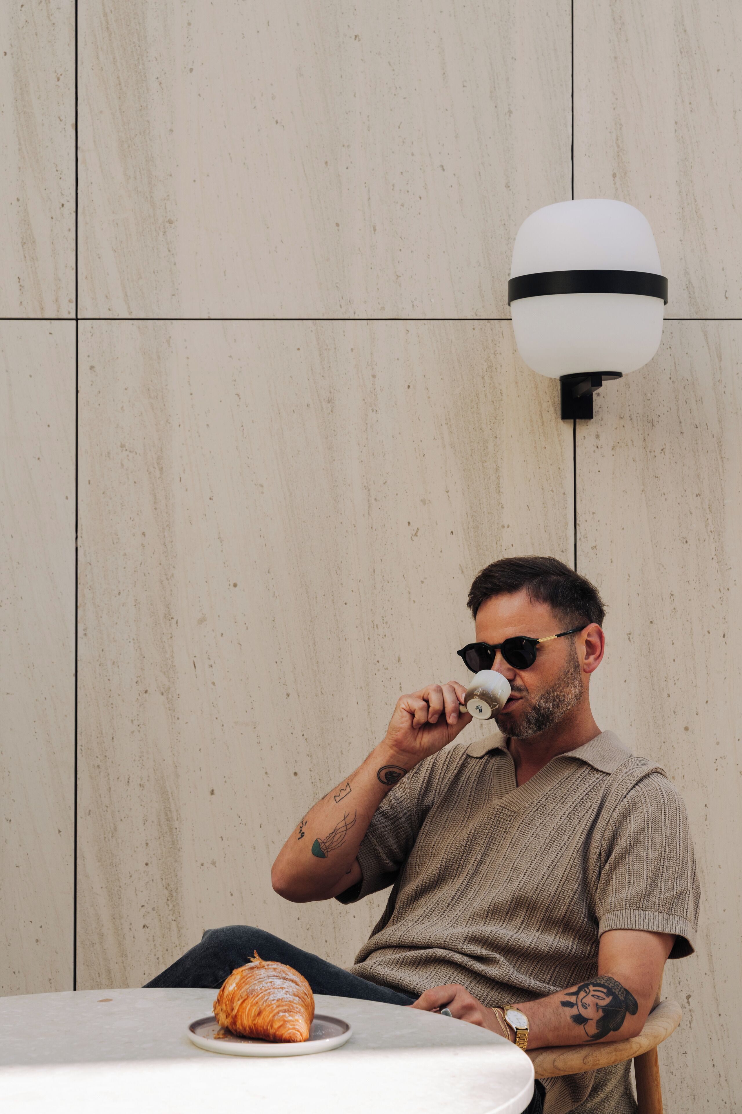 A man sitting at a table with a croissant, wearing a watch and a sweater, with a wall-mounted light fixture above.