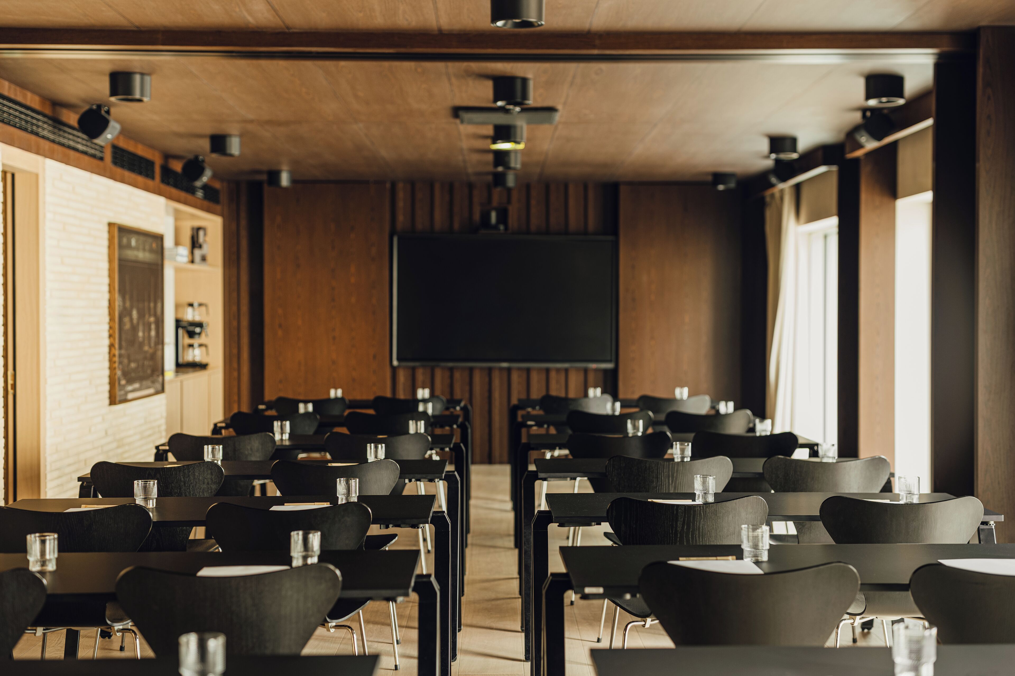 A conference room with rows of tables and chairs, a large screen, and wooden paneling on the walls.