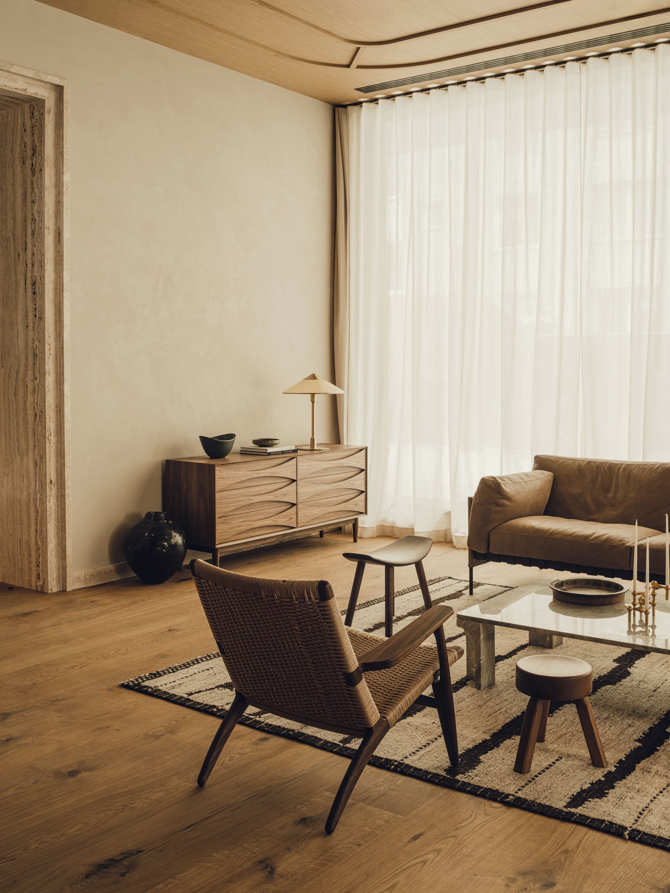 A living room with a wooden floor, a brown leather sofa, a woven chair, a glass coffee table, a small side table, a lamp, and a cabinet.