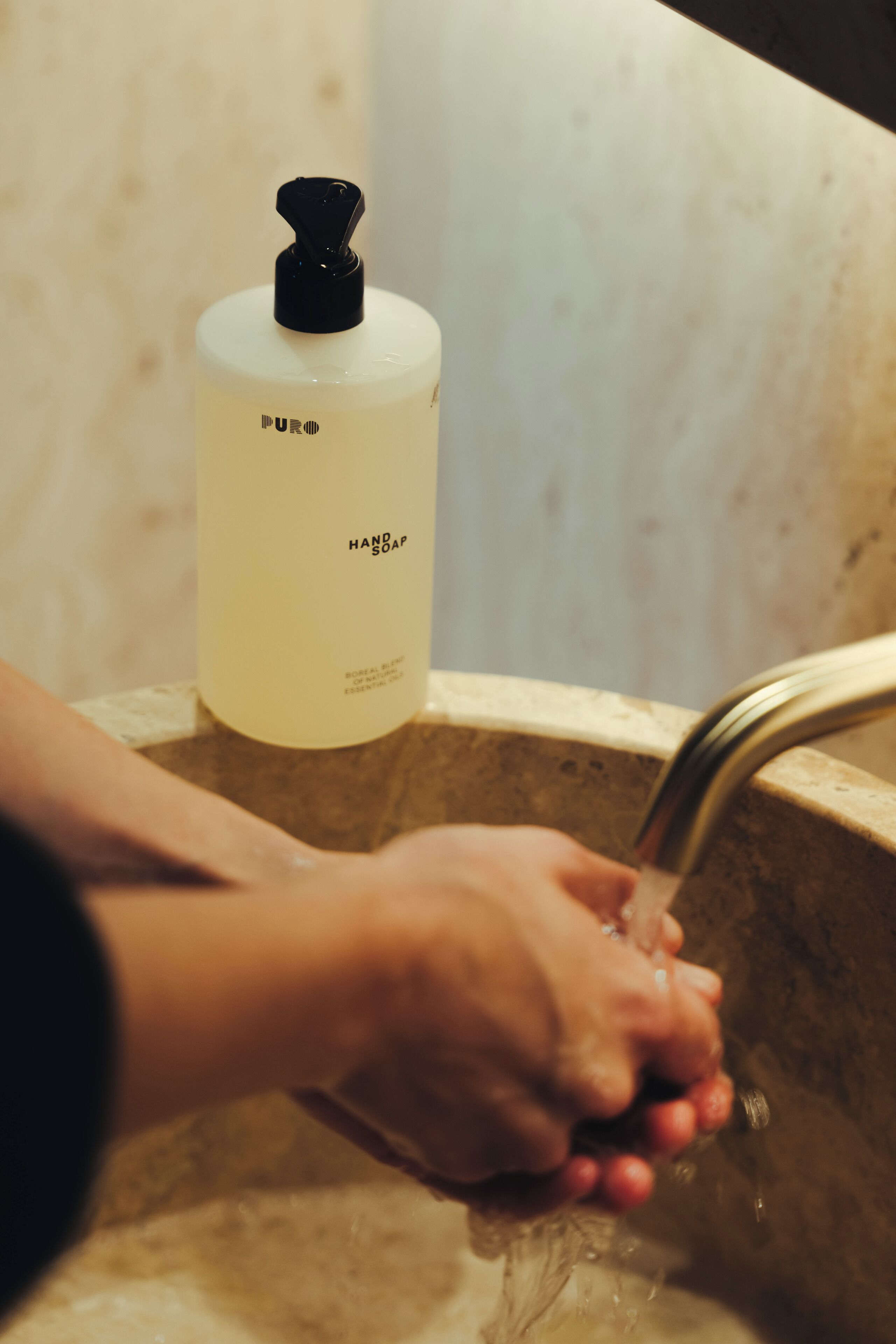 Een hand onder een stromende kraan met een fles handzeep met de tekst 'PURE HAND SOAP' op het aanrecht.