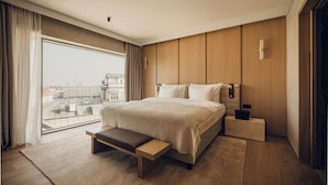Habitación de hotel moderna con una cama grande, un banco, una mesita de noche y vistas a la ciudad.